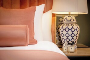A neatly made bed with a pink pillow, a white pillow, and a bedside table with a patterned lamp, a small vase, and two candles. - Hyde Hotel (Galway)