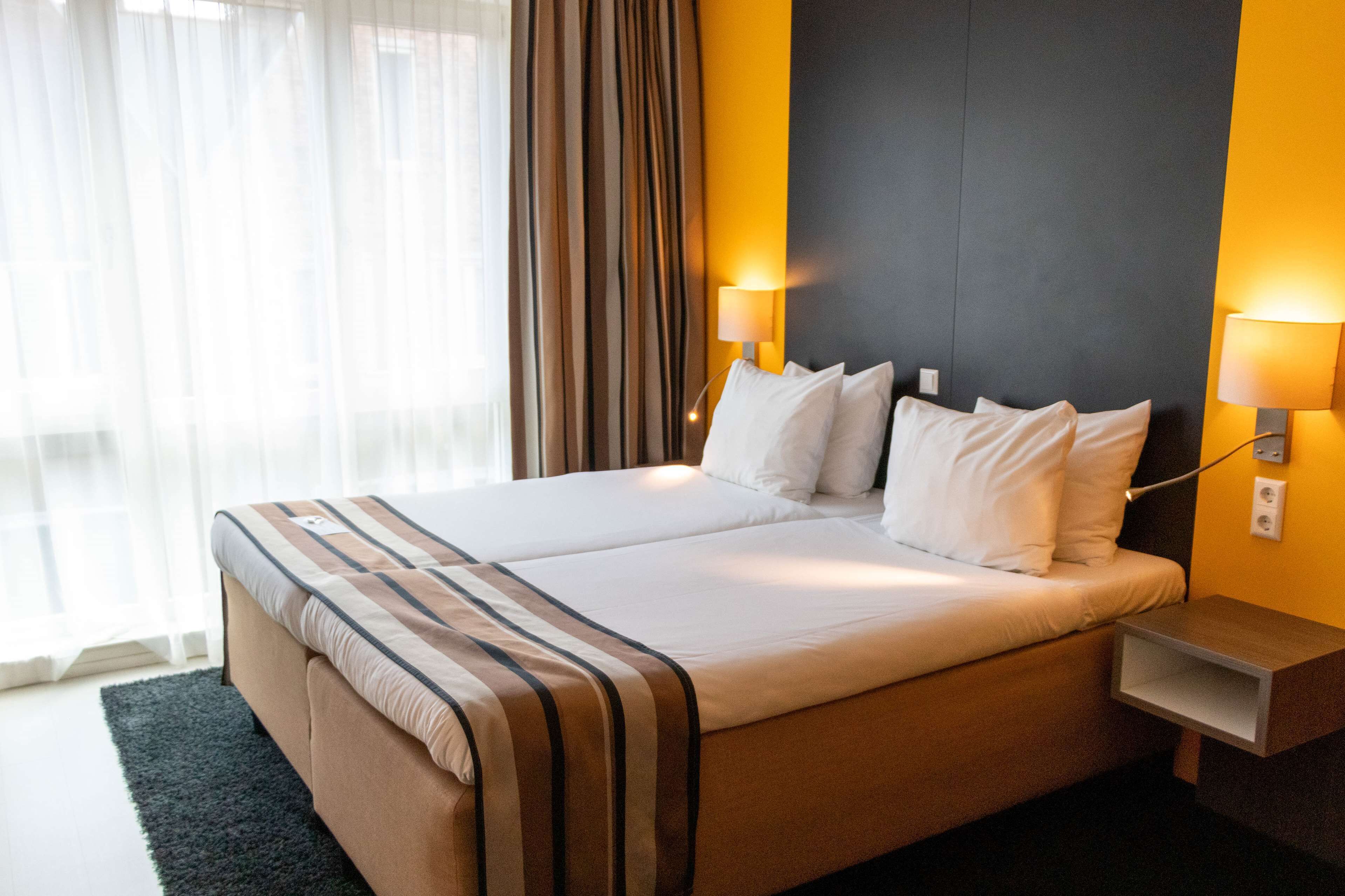 A double bed with a striped bedspread, two bedside lamps, a nightstand, and a view of a window with curtains.