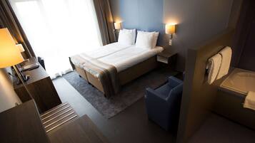 Habitación de hotel con cama, mesitas de noche, escritorio, silla, baño con bañera y ventana con cortinas.