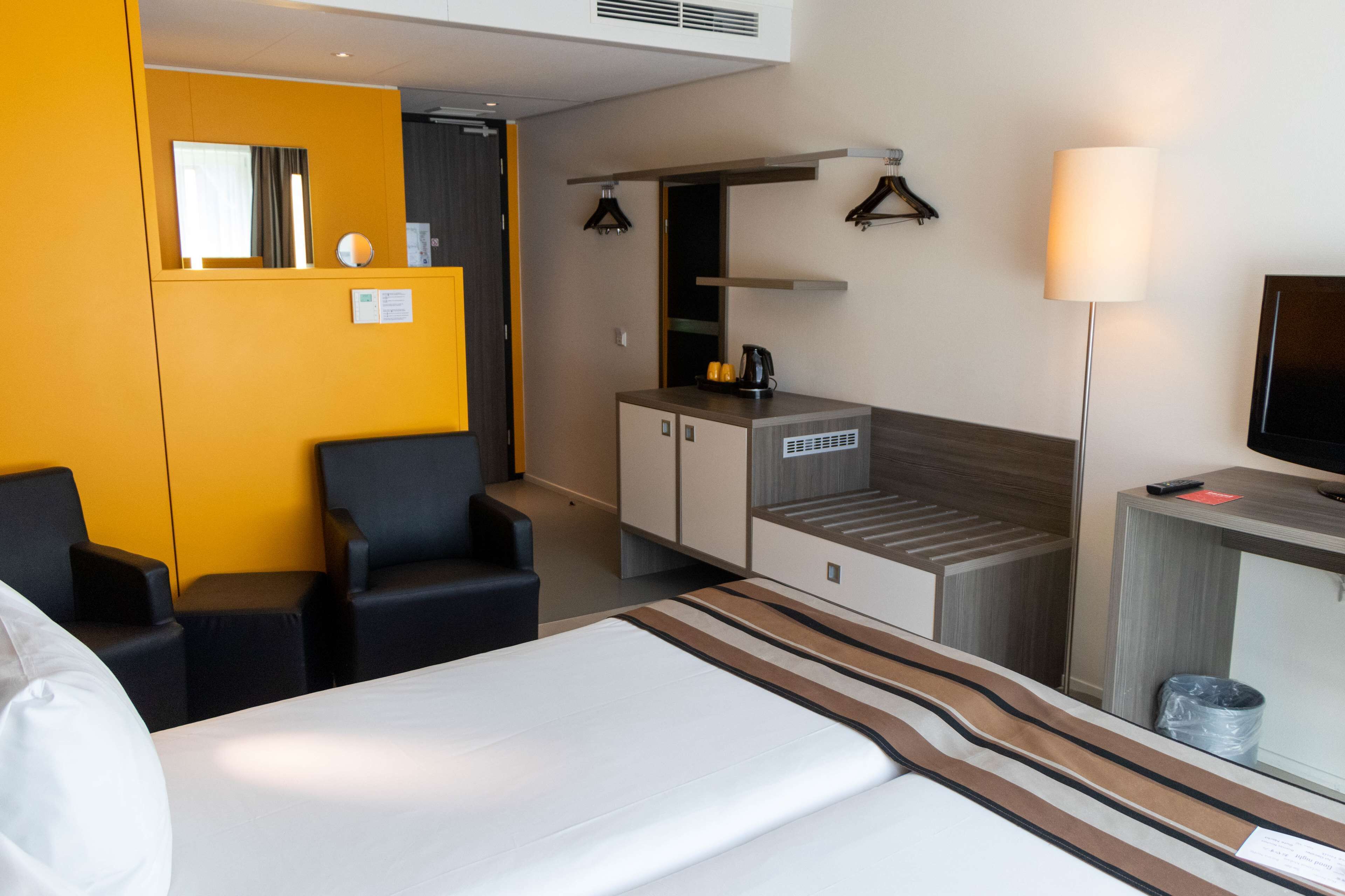 Una habitación de hotel con cama, escritorio, silla, kitchenette pequeña y televisión.