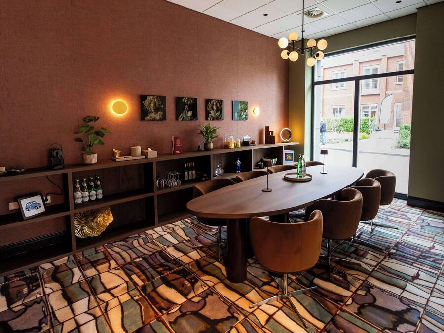 A modern meeting room with a large table, comfortable chairs, a bookshelf with various items, and a large window offering a view of the outside.
