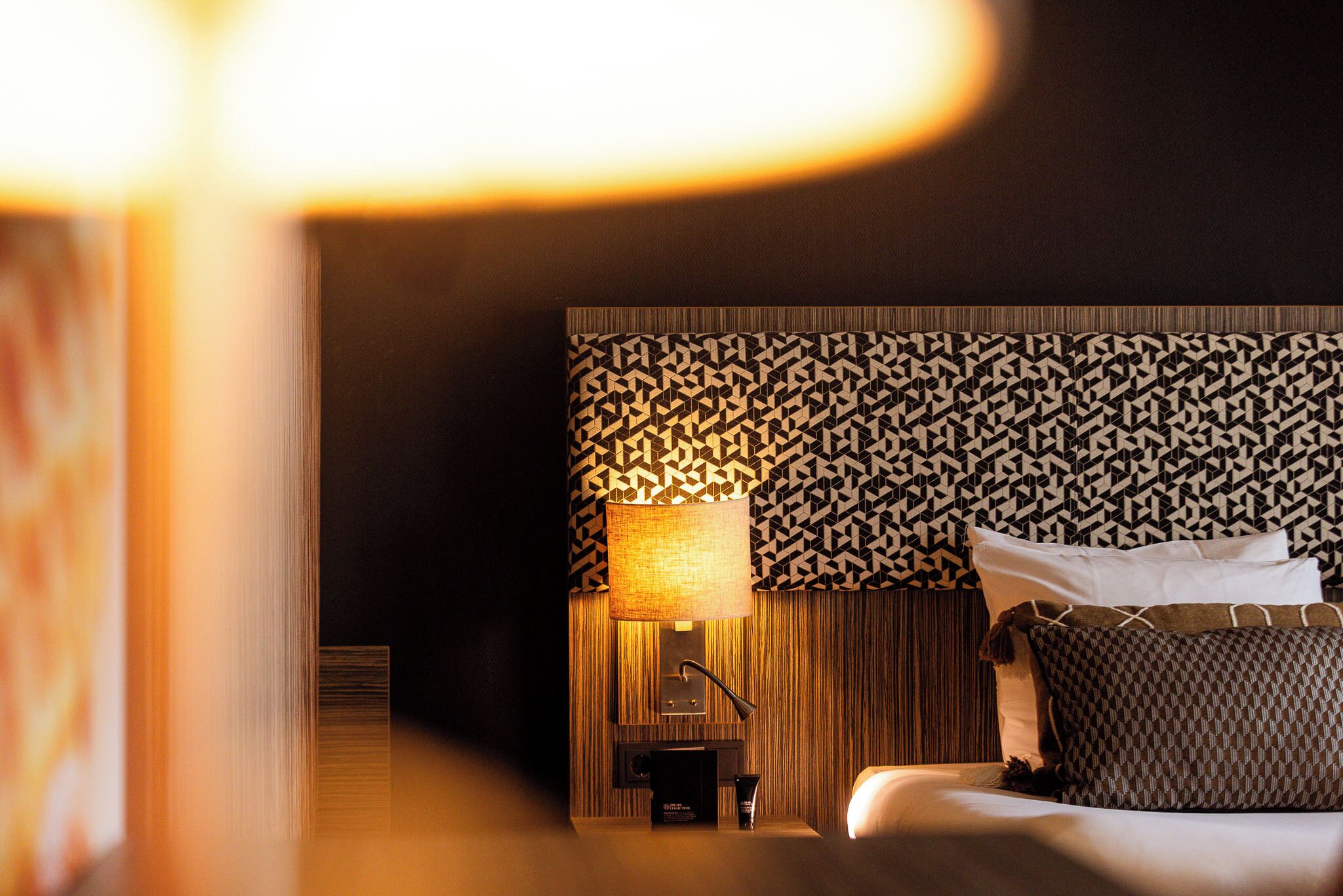 A hotel room with a patterned headboard, a bedside table with a lamp, and a bed with pillows.