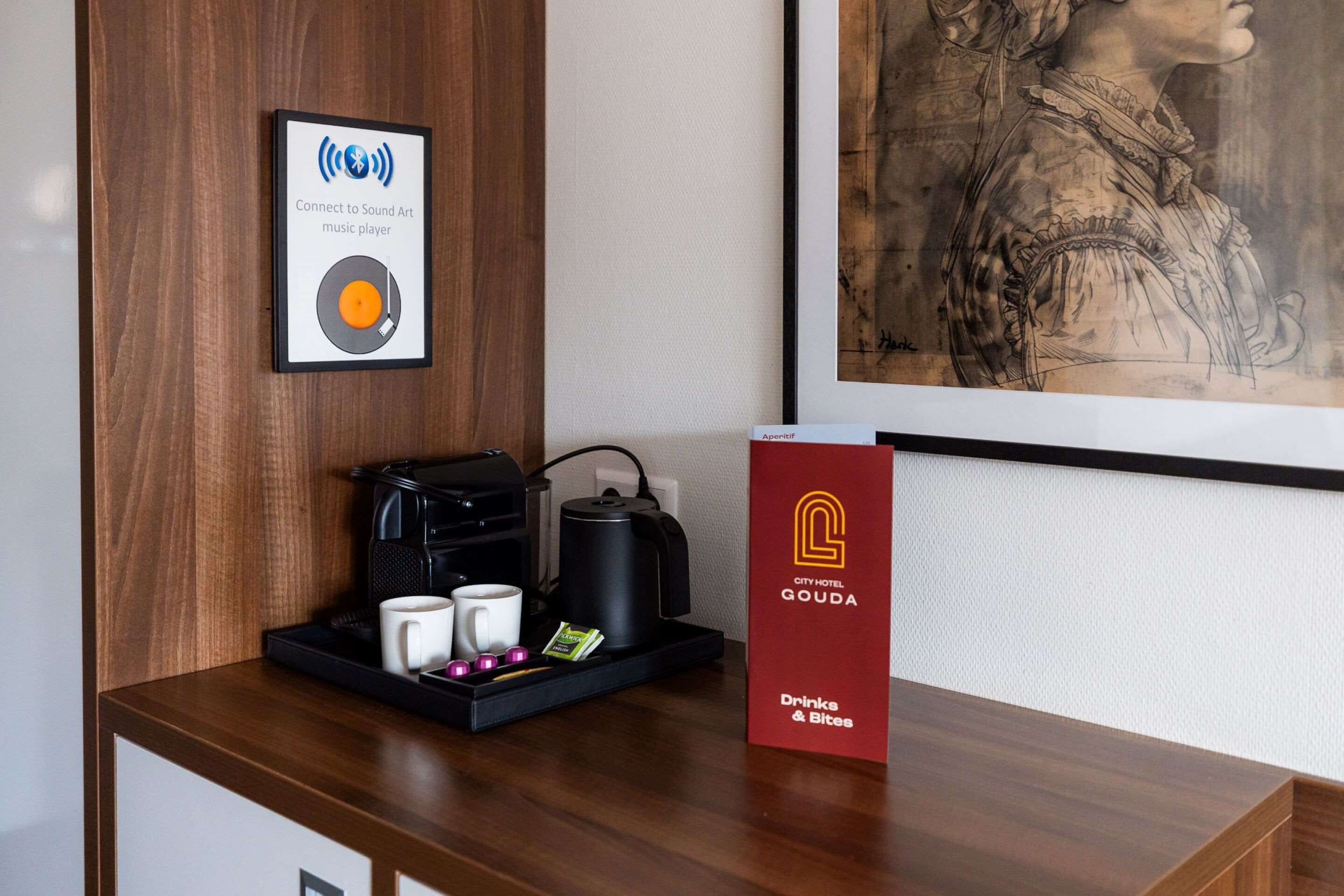 Uma área de recepção de hotel com uma máquina de café, xícaras, um tabuleiro, uma obra de arte emoldurada e uma caixa vermelha com a inscrição 