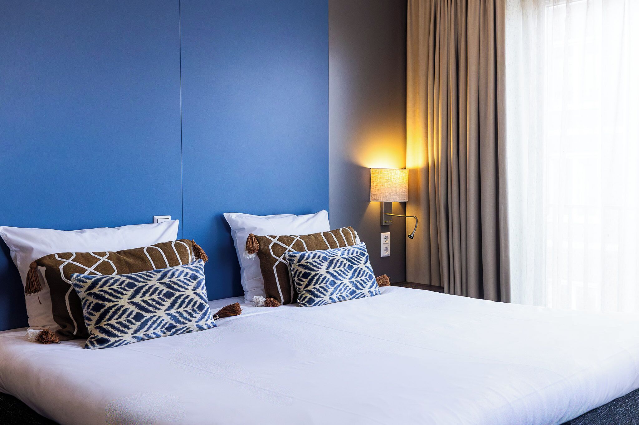 Una cama bien hecha con sábanas blancas y almohadas estampadas, una lámpara de pared y una ventana con cortinas.