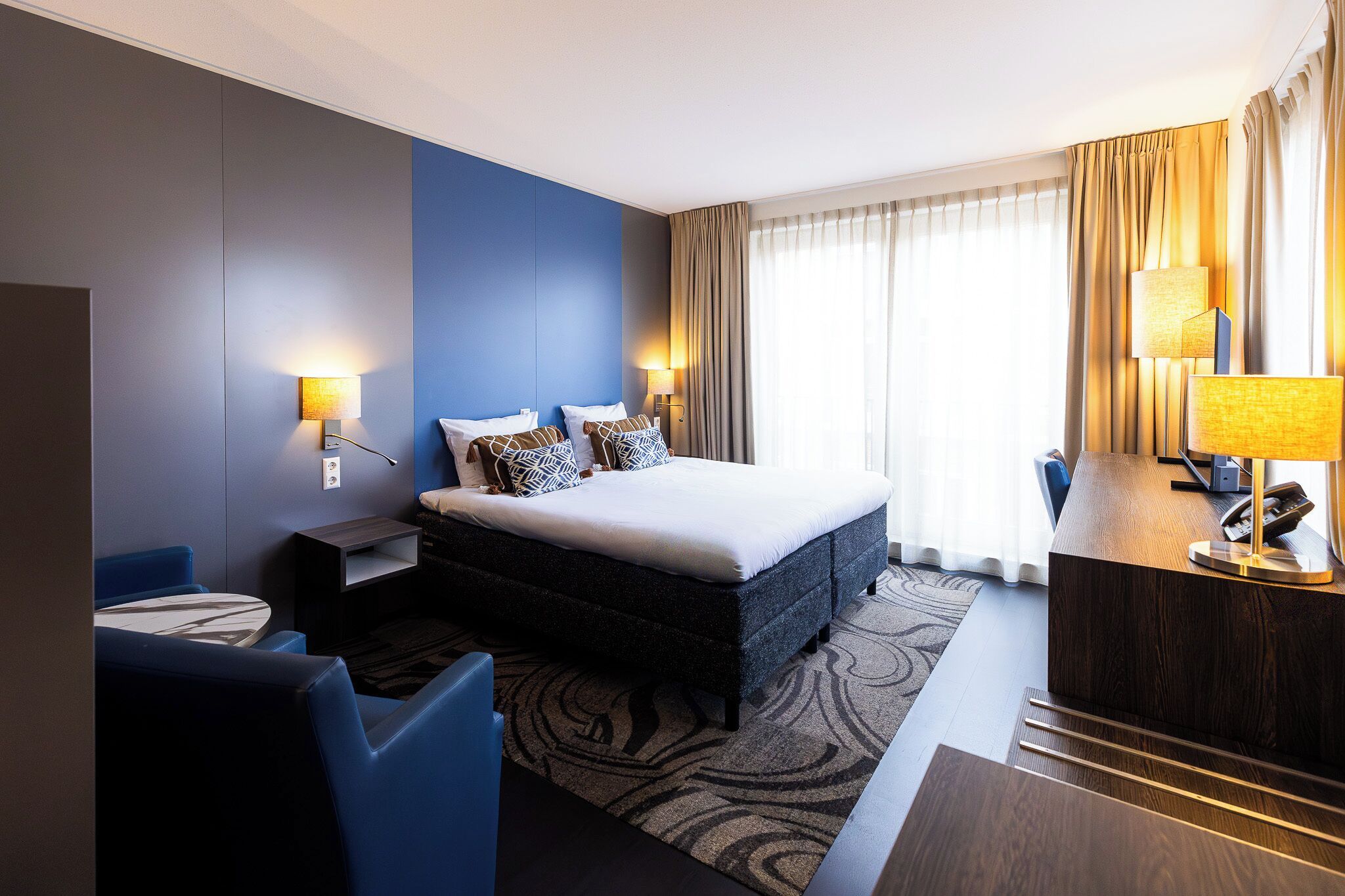 A hotel room with a large bed, two bedside tables with lamps, a desk with a lamp, and a view of the outdoors through the window.
