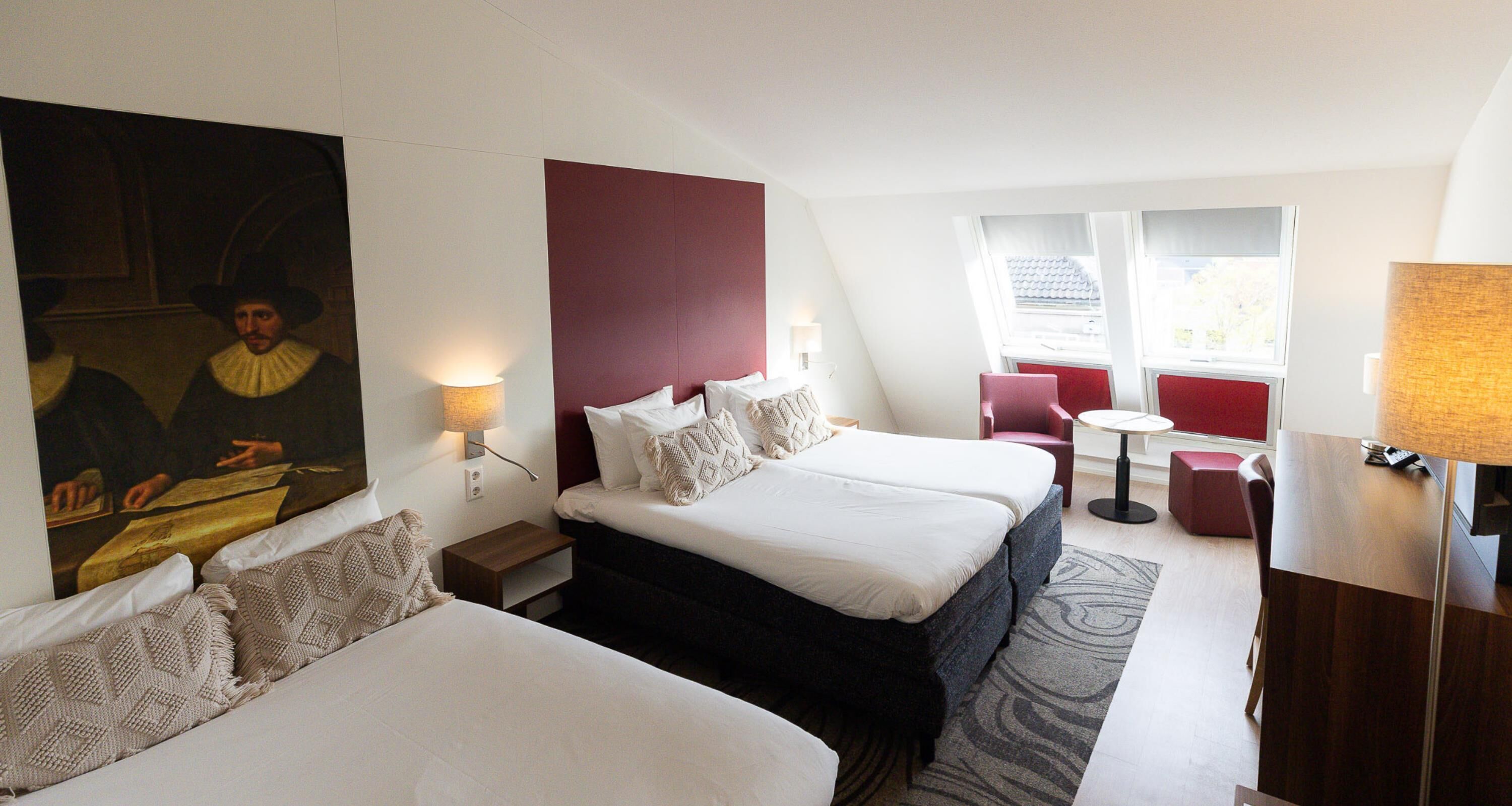 A hotel room with two beds, a red accent wall, a painting, a small table, and a red chair.