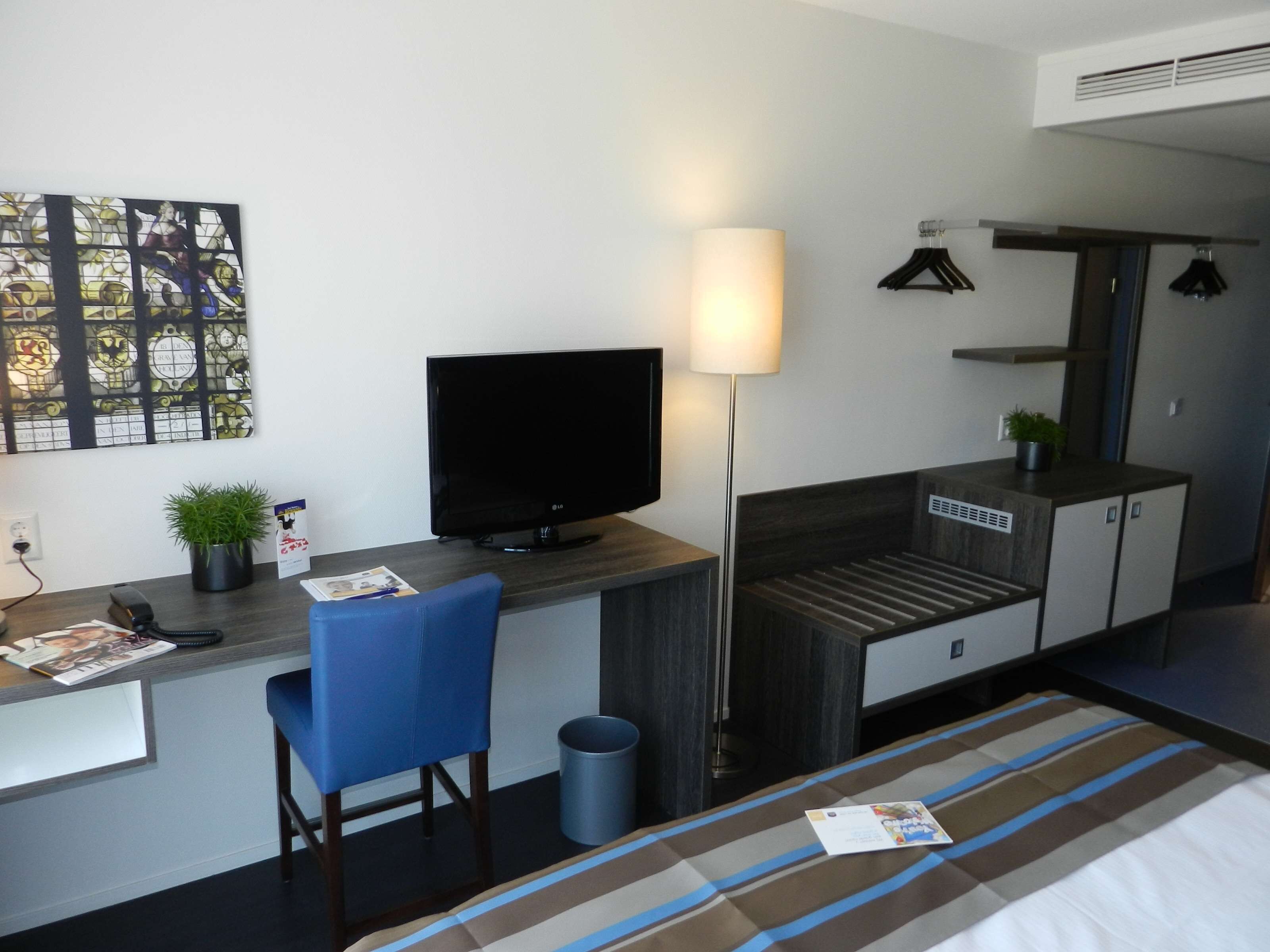 Een compacte hotelkamer met een bureau, stoel, televisie, bed en een kleine potplant.