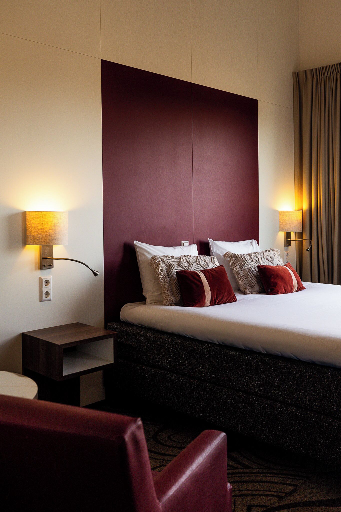 Een hotelkamer met een groot bed, twee bedlampen, een nachtkastje en een rode stoel.
