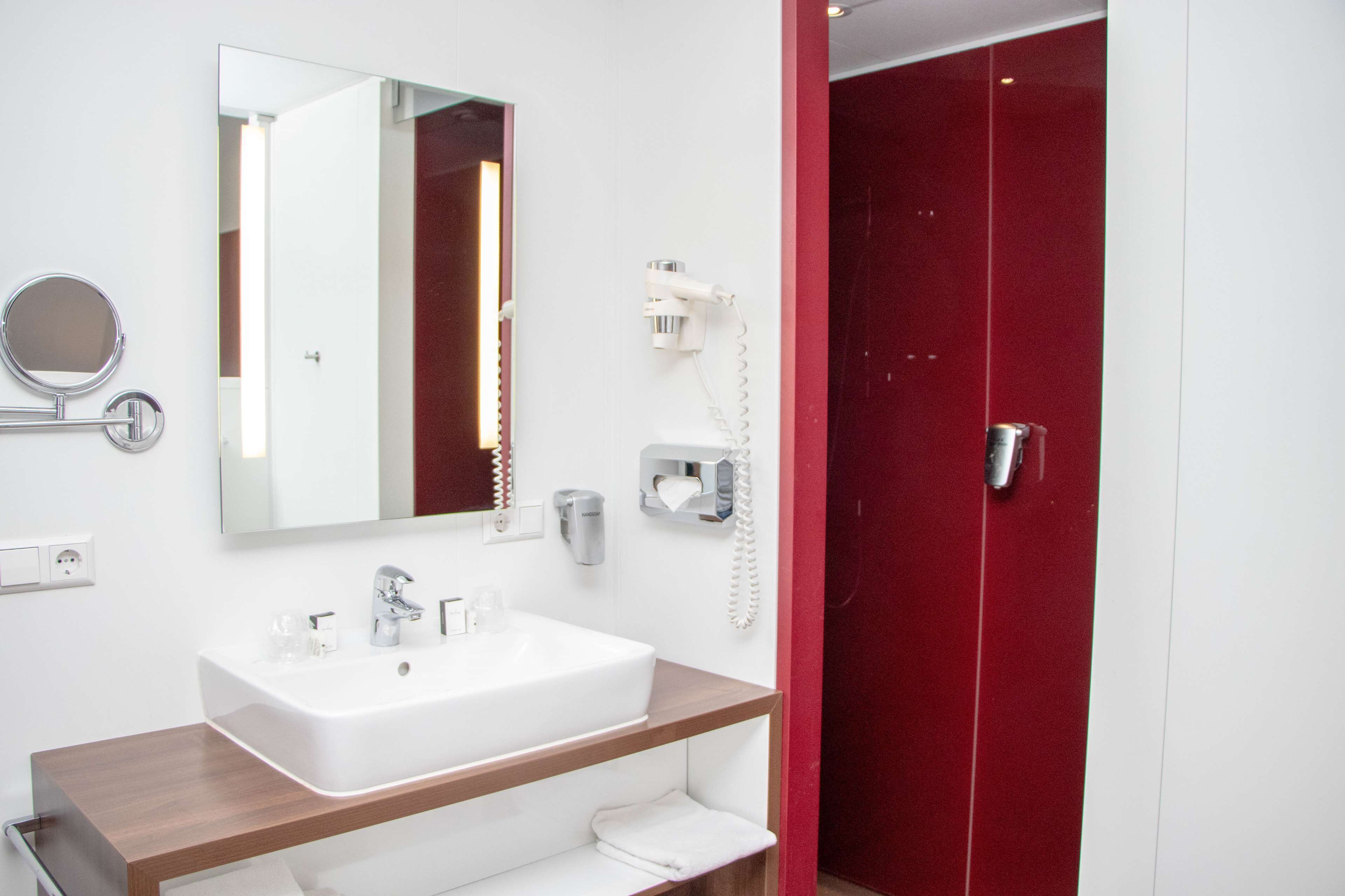 A modern bathroom with a white sink, a mirror, a hairdryer, and a red door.