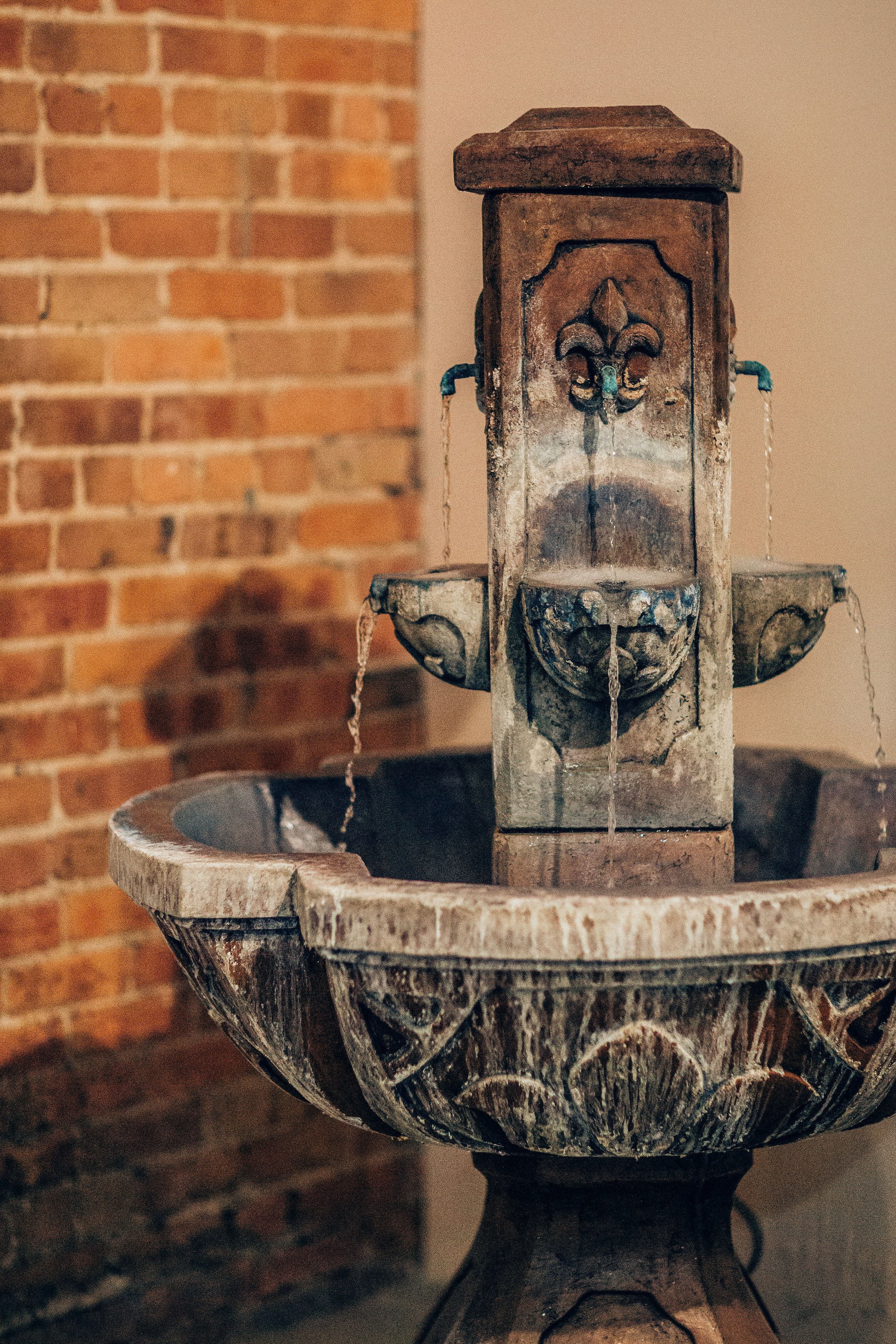 A rustic stone water fountain with a decorative basin, set against a brick wall.