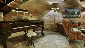 Een kamer met een vleugelpiano, een leren bank, een gestreept fauteuil, een vloerlamp en een schilderij op de bakstenen muur.