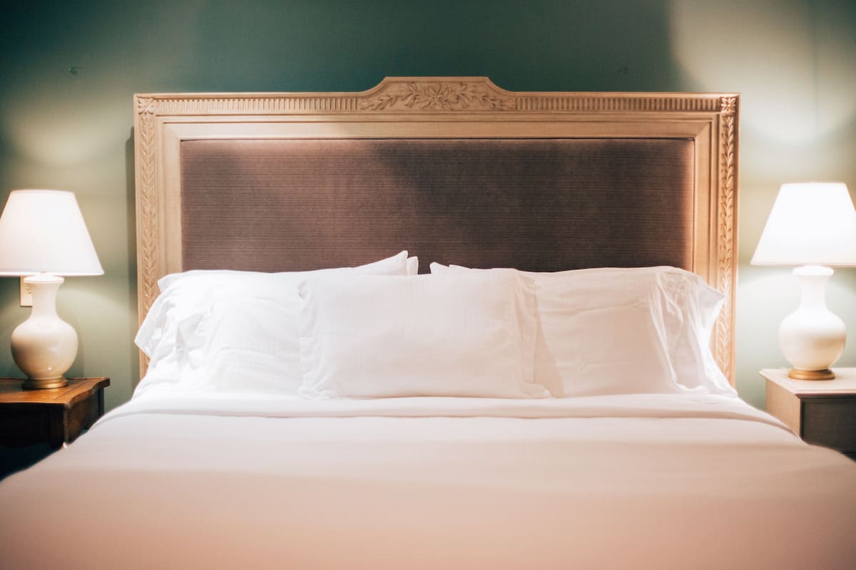 Een net opgemaakt bed met witte lakens en een houten hoofdbord, met aan weerszijden nachtkastjes en bijpassende bedlampen.