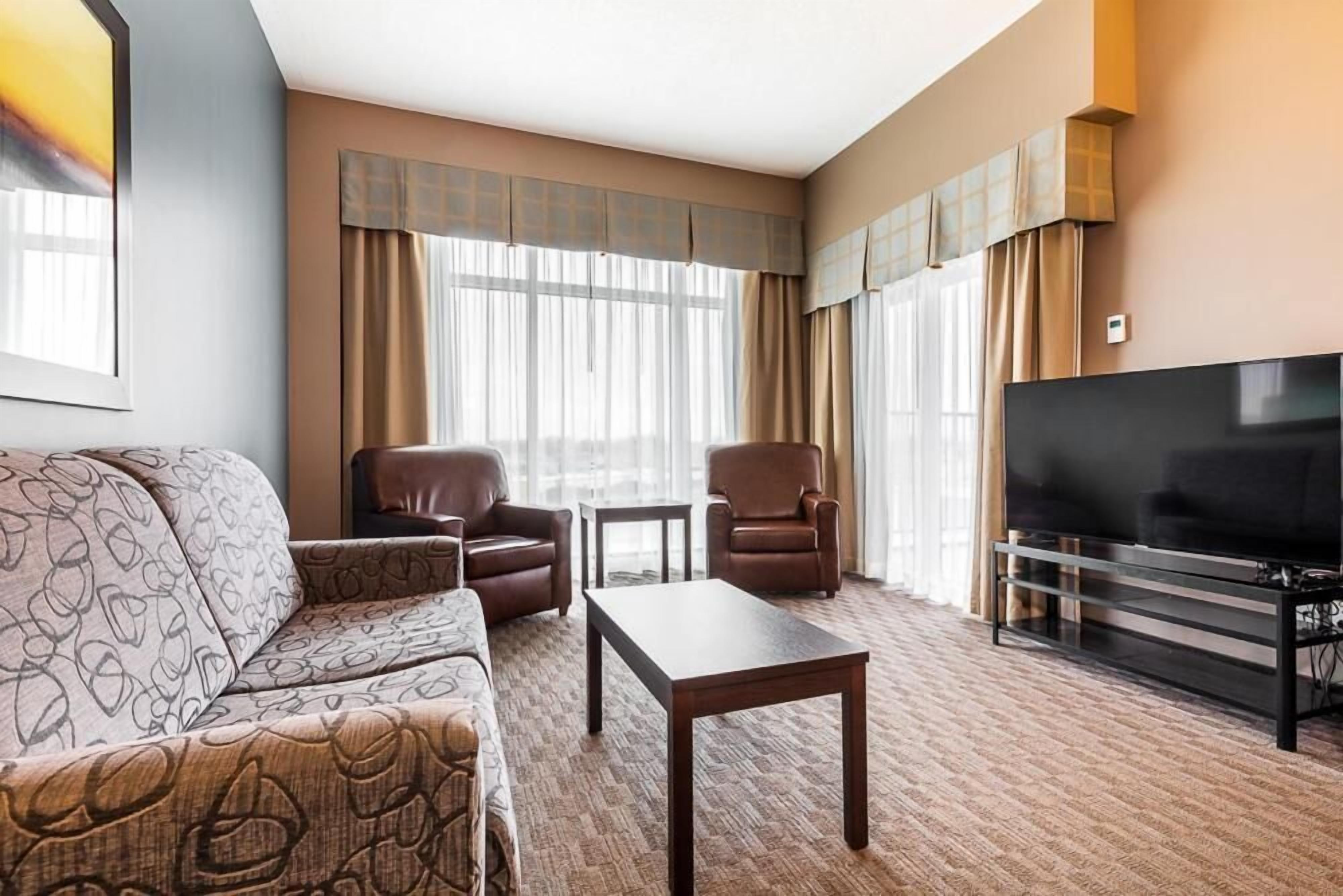 A hotel room with a sofa, two armchairs, a coffee table, a TV, and large windows with curtains.