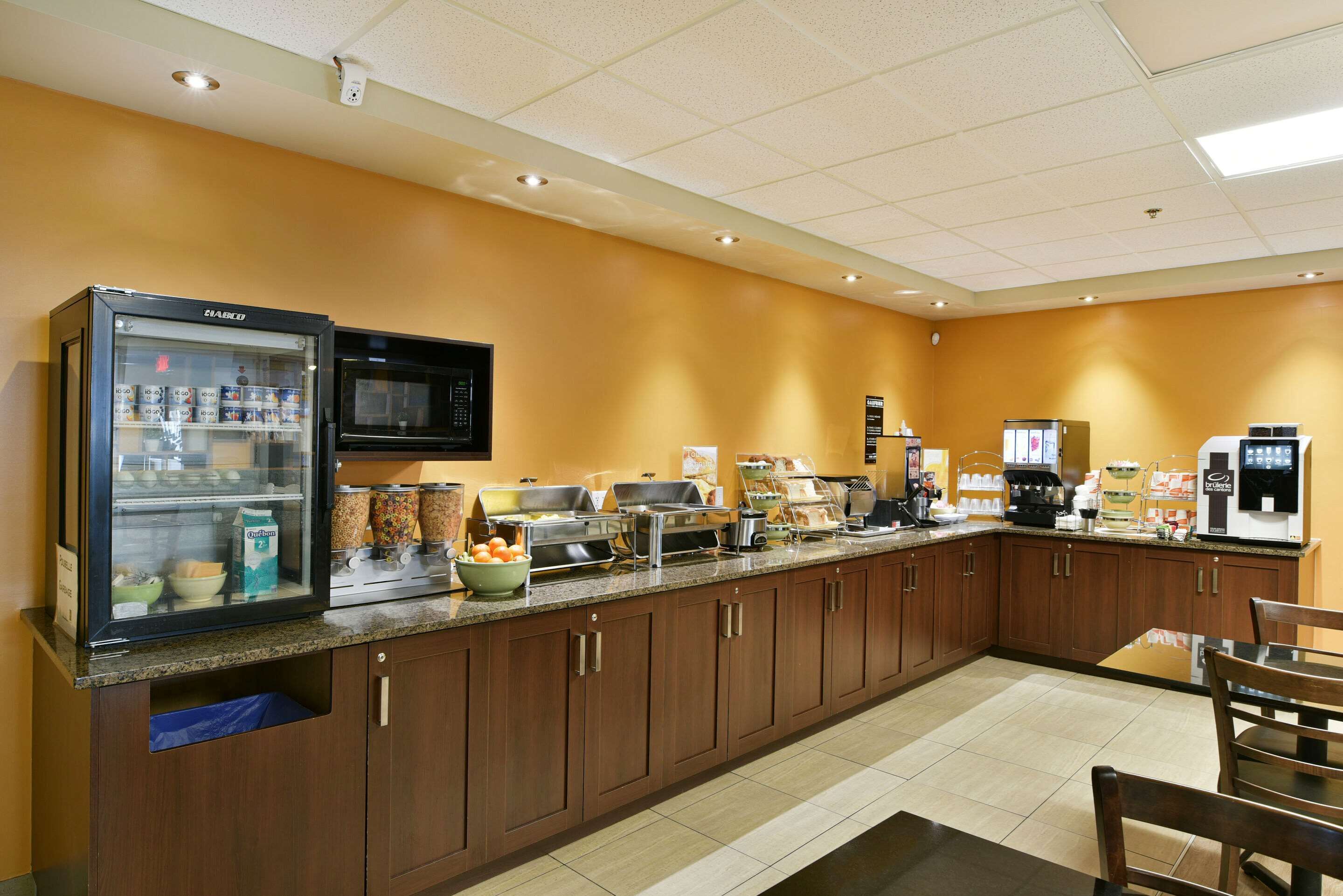 A hotel kitchen with a self-service breakfast bar, a refrigerator, microwave, and various food items.