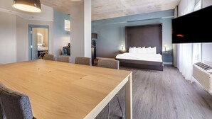 A modern hotel room with a wooden dining table, two pendant lights, a large bed with a headboard, a flat-screen TV, and a large window with curtains.