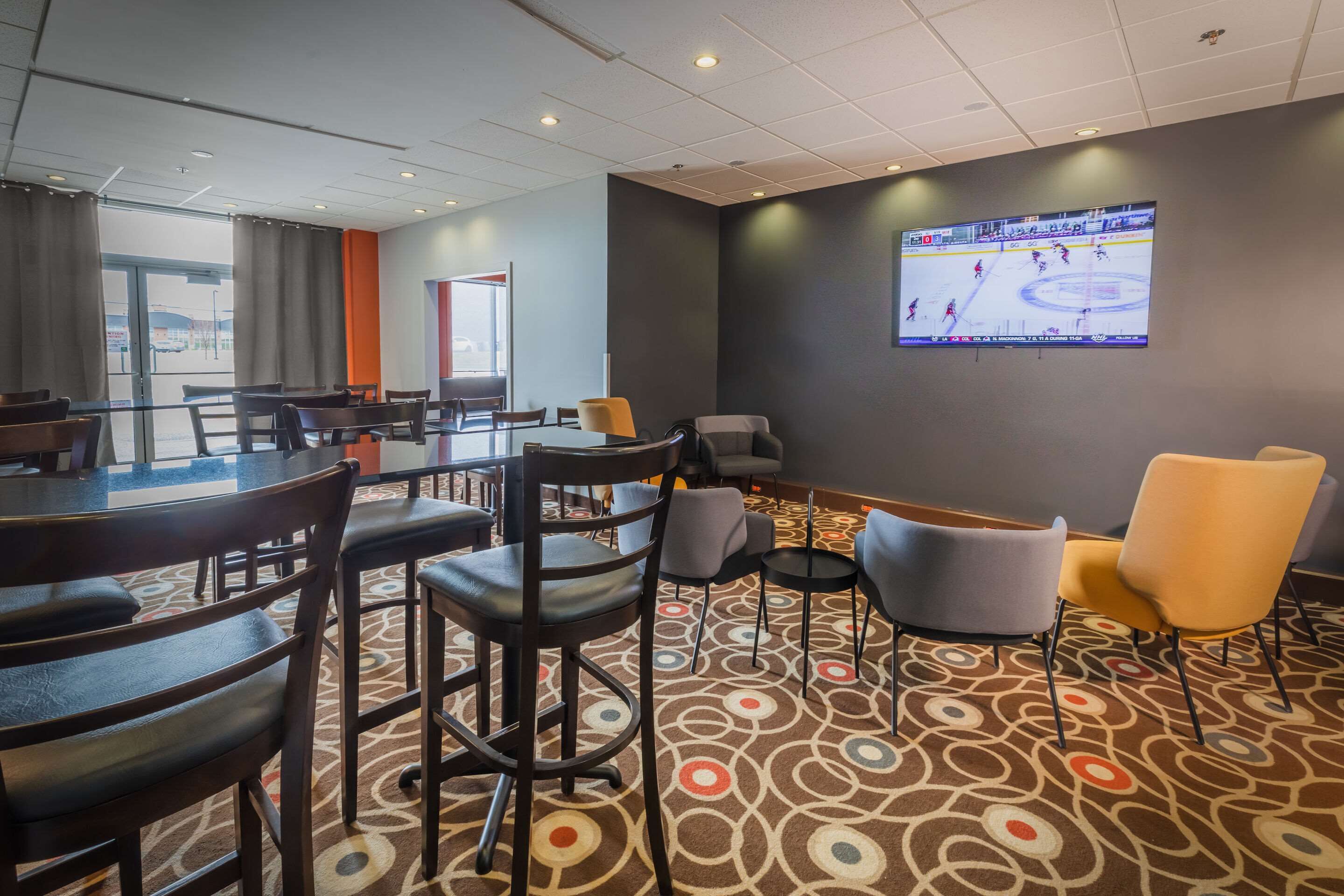 Een hotel lobby met een groot scherm waarop een ijshockeywedstrijd wordt vertoond, diverse zithoeken met tafels en stoelen, en een kamerbreed tapijt met een patroon.