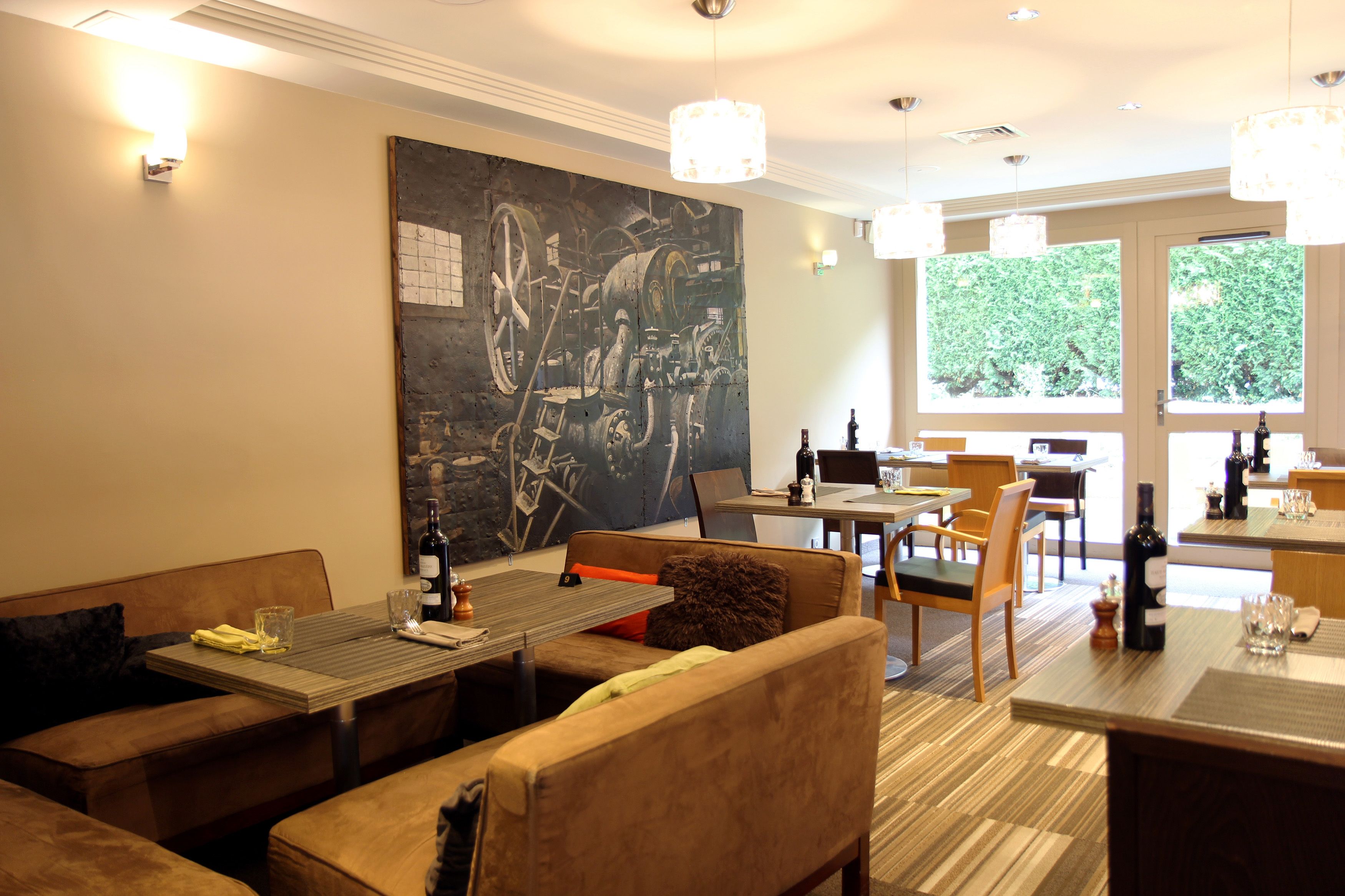 Un restaurant au décor moderne, avec un grand tableau abstrait accroché au mur, de nombreuses tables et chaises, et une vue sur la verdure par les fenêtres.