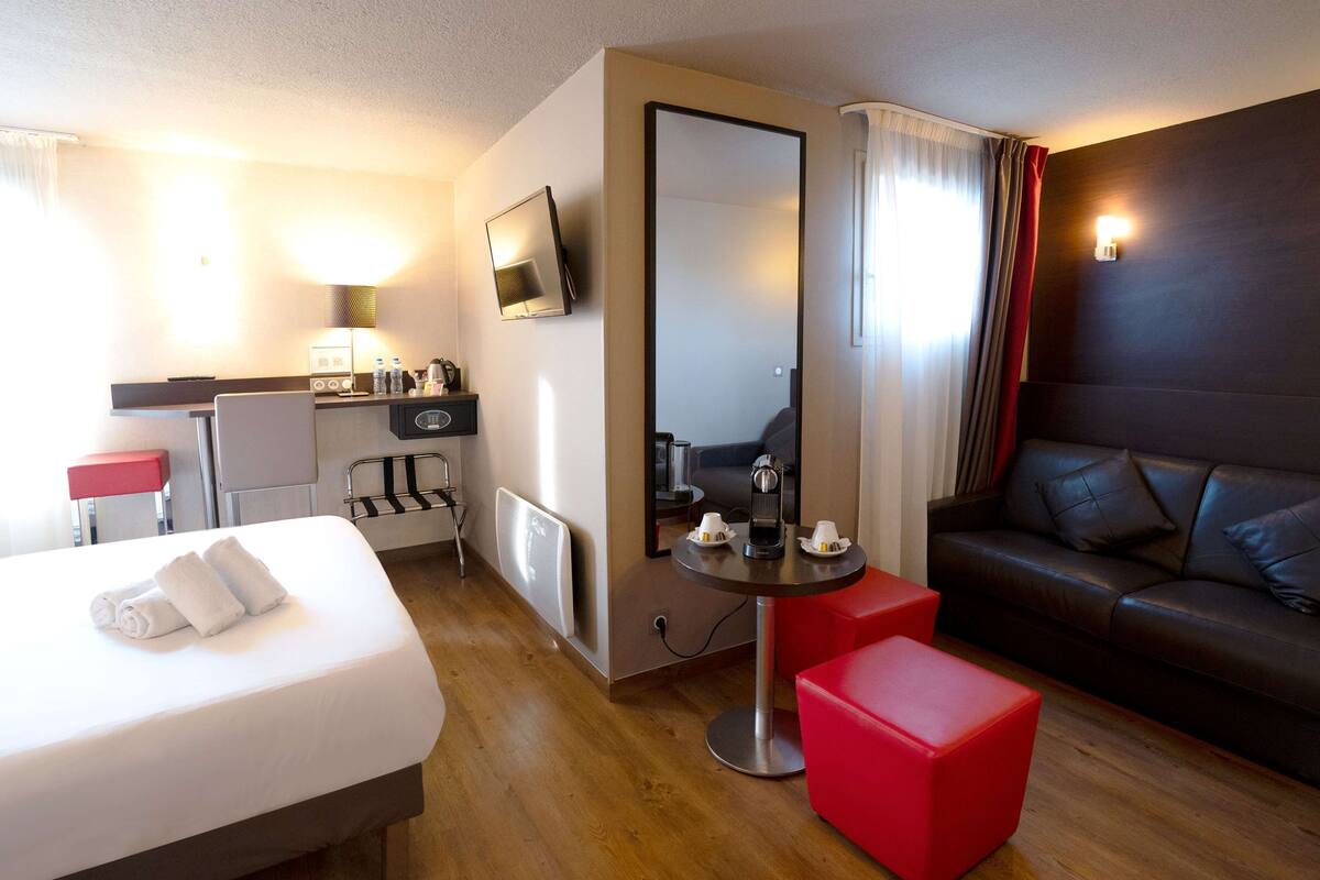 Une chambre d’hôtel comprenant un lit, un pouf rouge, un canapé, une petite table, un miroir, une télévision, un bureau avec une chaise et une kitchenette.