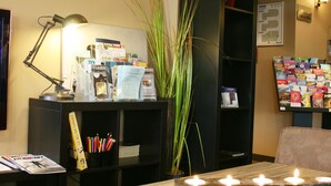 A hotel reception area with a desk, computer, lamp, and a bookshelf with magazines. - Best Western l'Atelier 117 (Maubeuge)