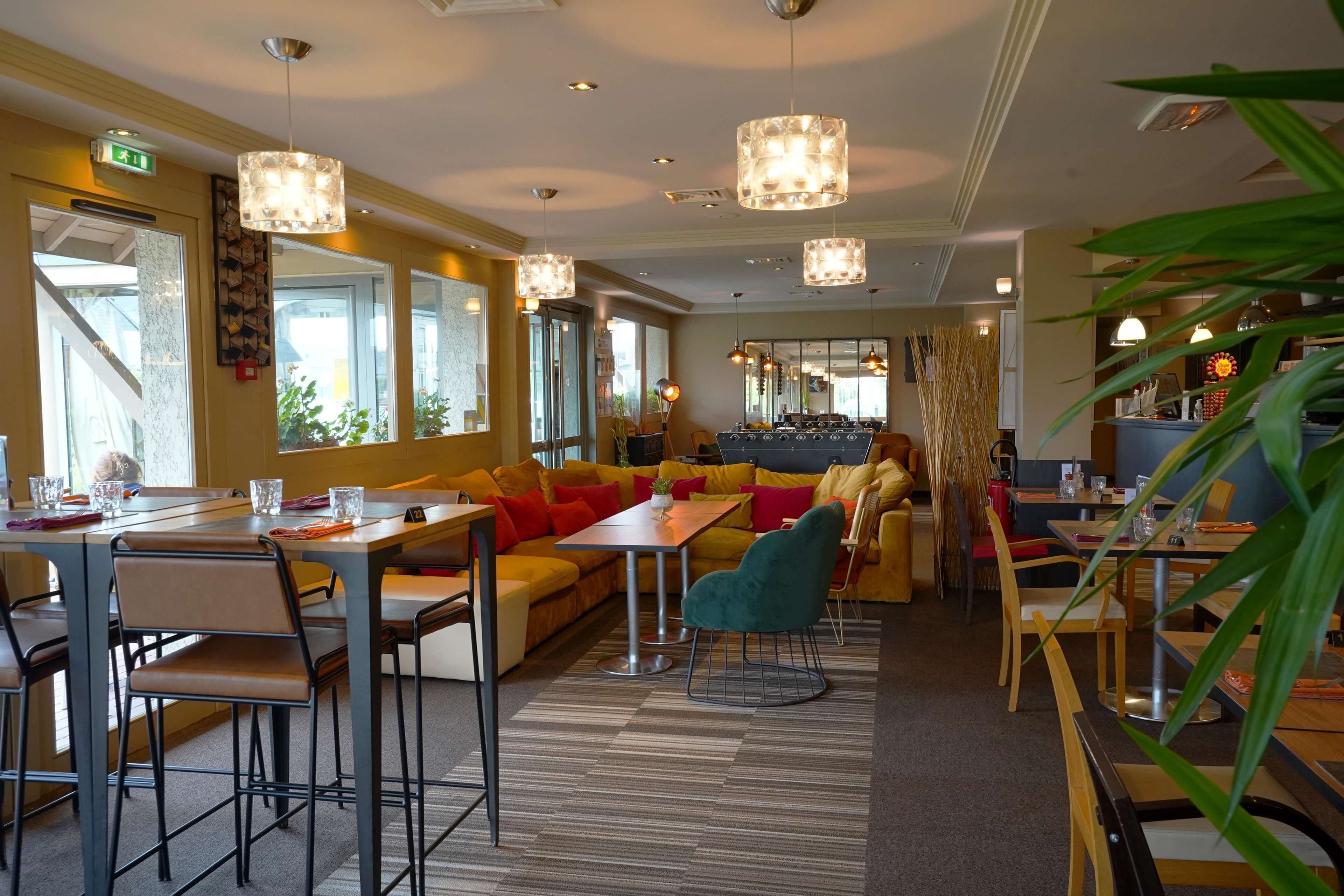 En moderne hotellobby med forskellige siddeområder, heriblandt en sofasætning, et spisebord og et barområde. Flere hængende pendellamper i loftet, og rummet er godt oplyst af naturligt lys fra vinduerne.