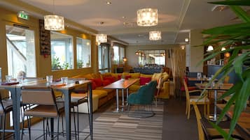 En moderne hotellobby med forskellige siddeområder, heriblandt en sofasætning, et spisebord og et barområde. Flere hængende pendellamper i loftet, og rummet er godt oplyst af naturligt lys fra vinduerne.