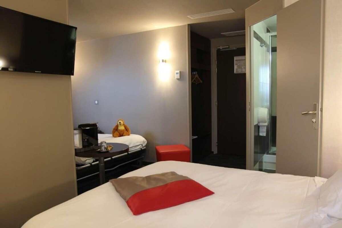 A hotel room with a bed, a television mounted on the wall, a small table with a coffee maker, and a red stool.