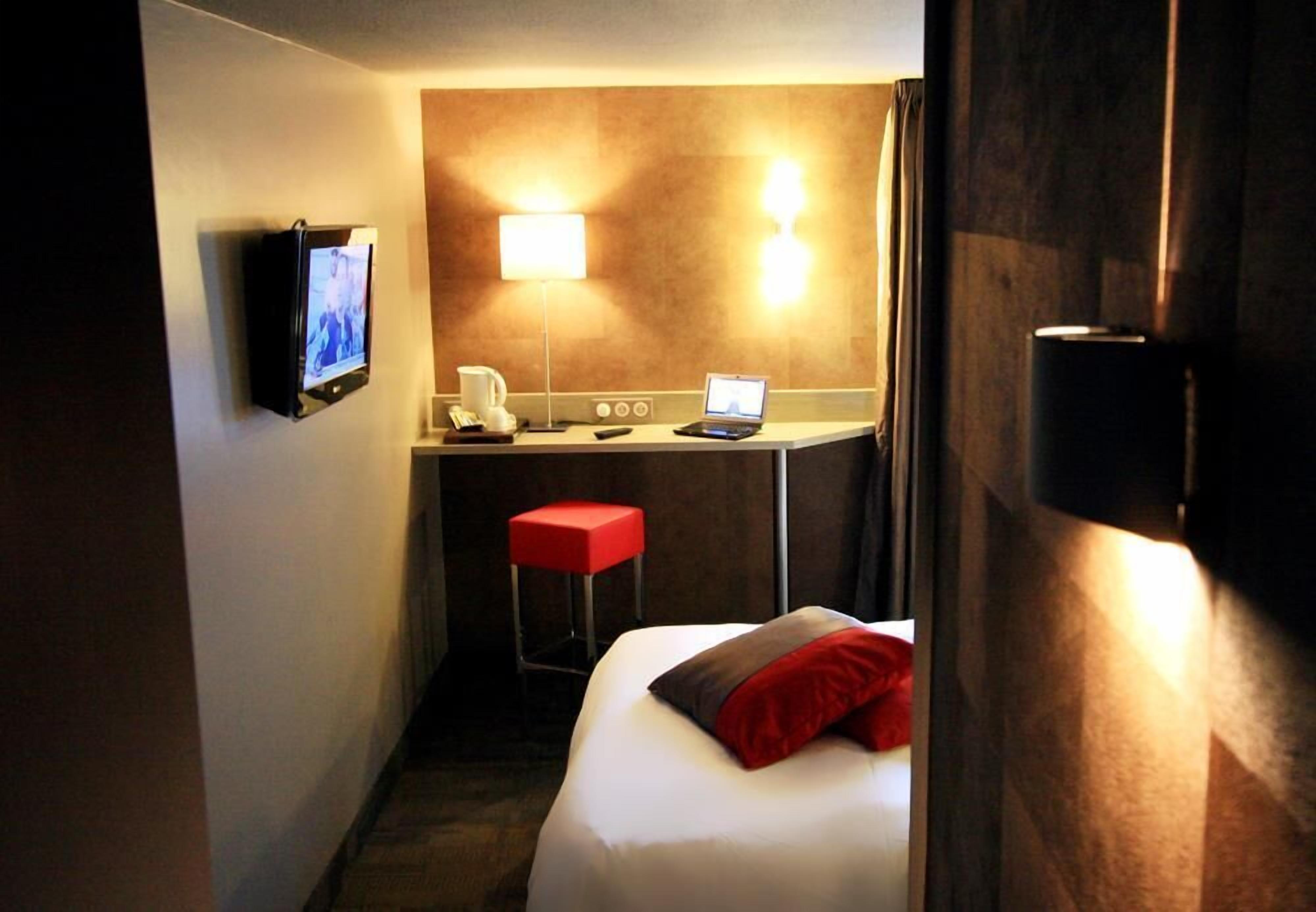 A hotel room with a bed, a desk with a laptop, a TV, and a red stool.