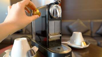 A hand holding a coffee pod, a coffee machine, and two white cups on a table.