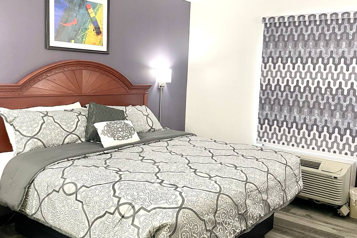 Un lit bien fait, avec une literie à motifs, une tête de lit, une œuvre d’art encadrée au mur et une fenêtre agrémentée de rideaux à motifs.