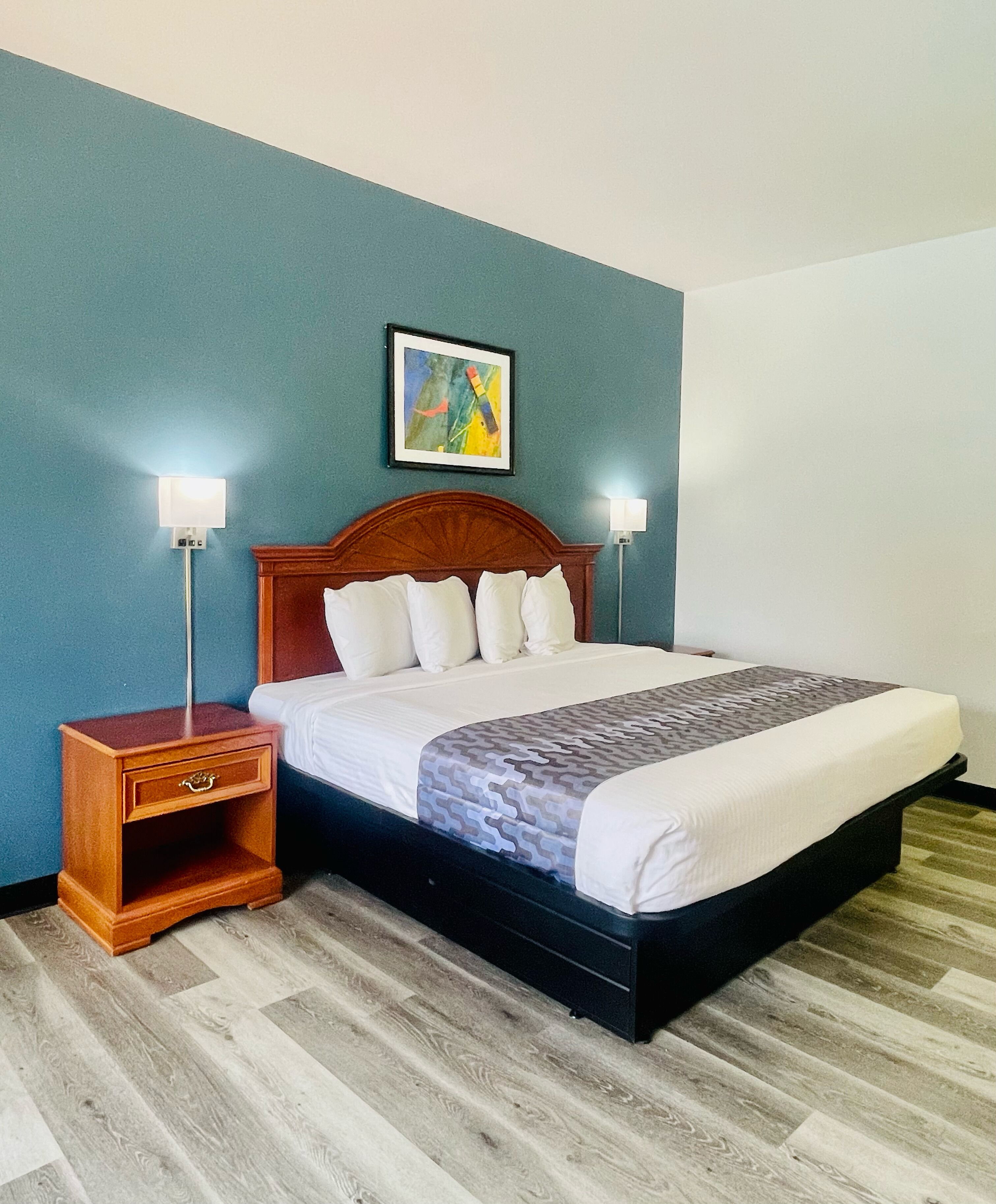 A neatly made bed with a patterned bedspread, a wooden nightstand with a drawer, two wall-mounted lamps, and a framed abstract painting above the bed.
