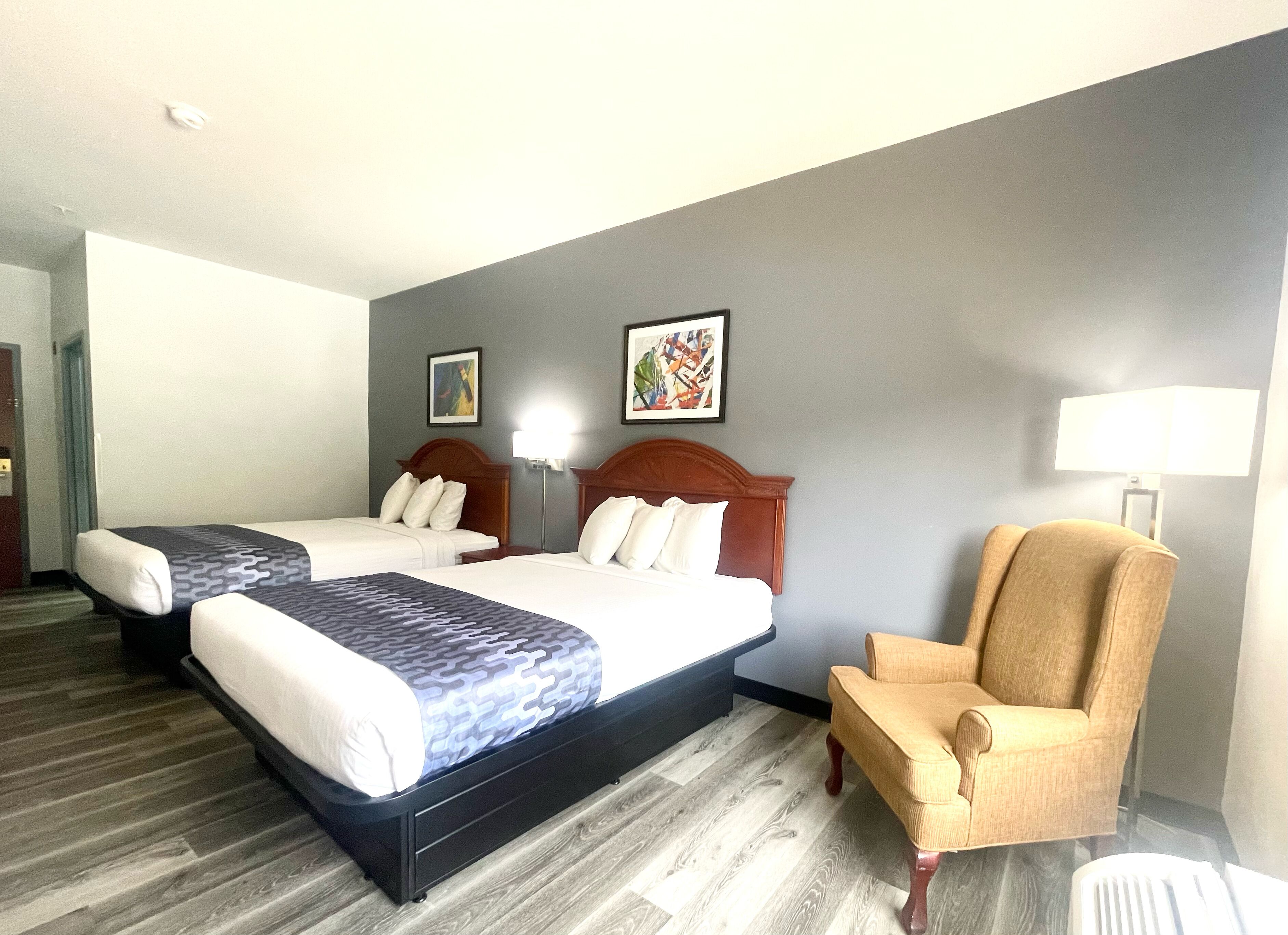 A hotel room with two beds, a chair, a lamp, and artwork on the walls.