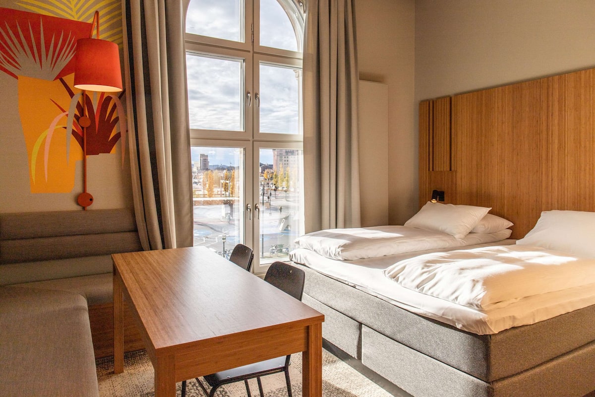 Una camera d'albergo con un letto, un tavolo in legno, una poltrona e un'ampia finestra con vista sulla città.