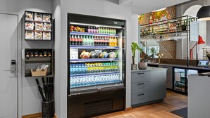 A modern hotel lobby with a large beverage dispenser, snack vending machine, and a small bar area with a plant and a red lamp. - Comfort Hotel Grand Central (Oslo)