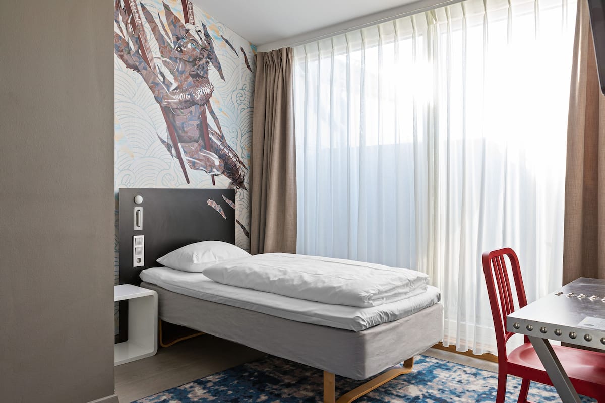 Camera d'albergo con un letto, una scrivania, una sedia e un'ampia finestra con tende.