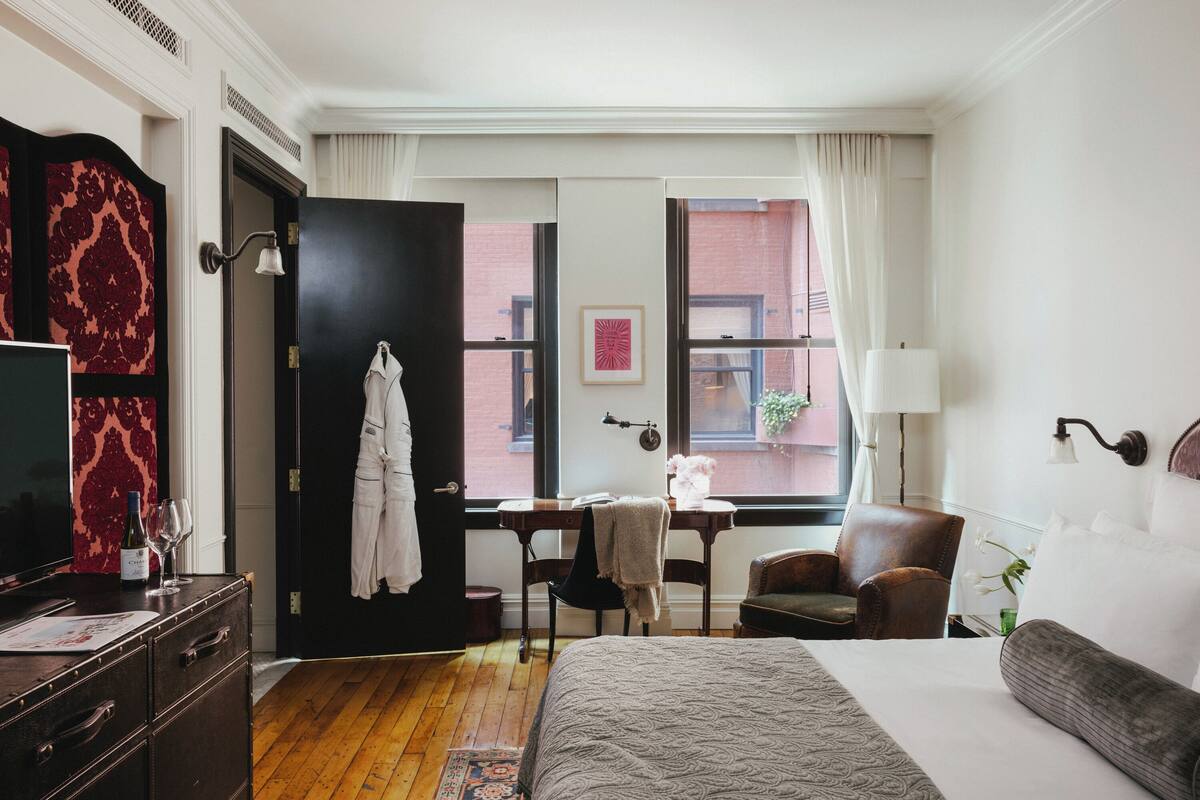 Camera d'albergo con un letto, una scrivania, una sedia, una TV e una finestra con vista su un edificio.