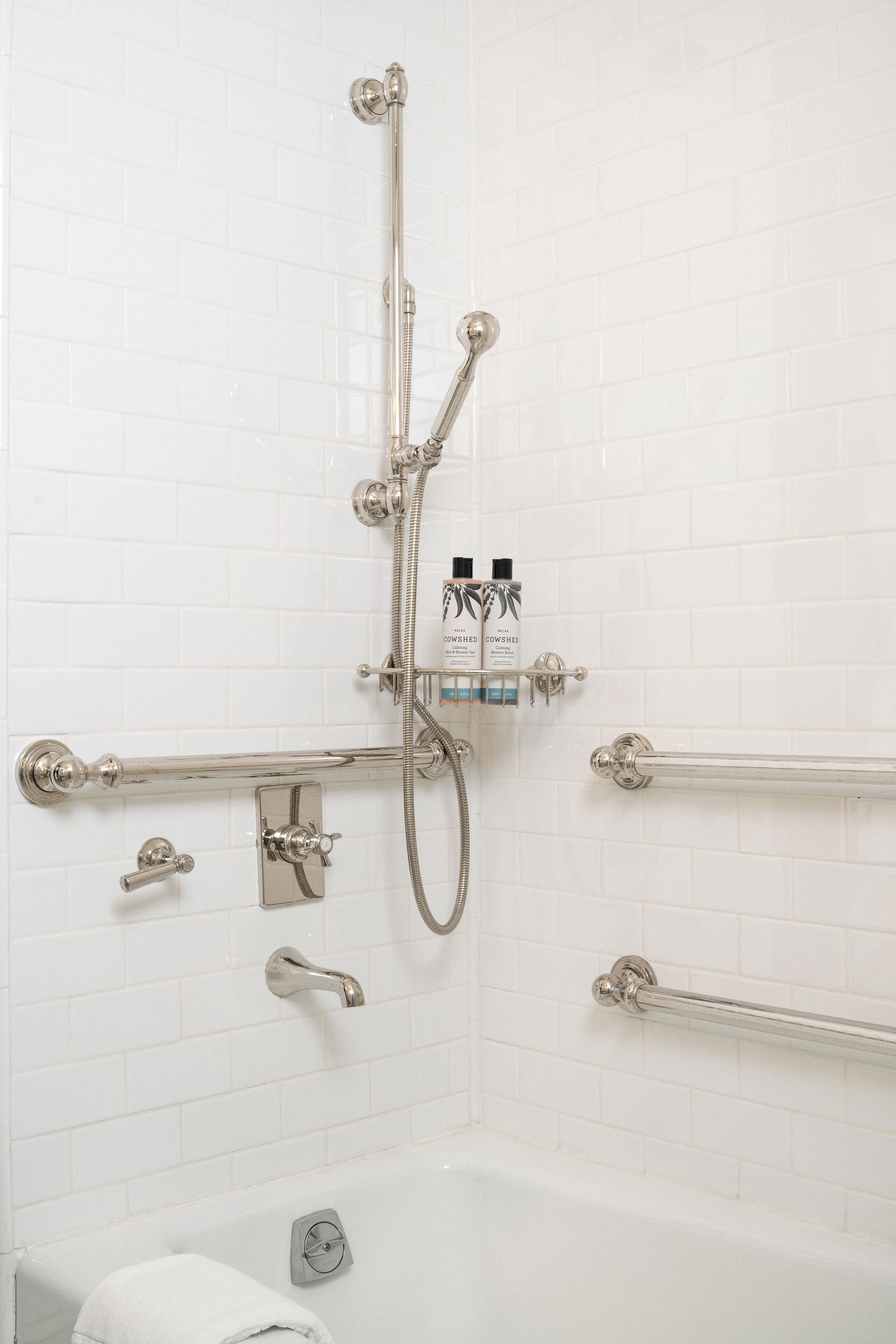 A bathroom with white tiled walls, a bathtub, a shower with a handheld showerhead, and a grab bar.