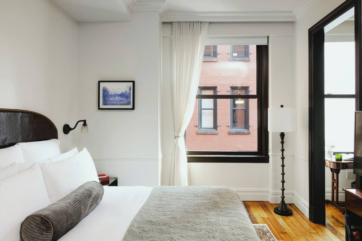 Una camera d'albergo con un letto, una finestra vista su un edificio in mattoni, un quadro al muro e una lampada da terra.