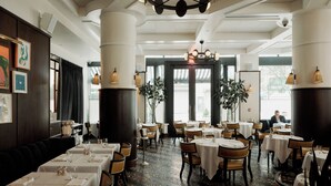 A restaurant with round tables and chairs, a chandelier, and large windows. - The Ned NoMad (New York)