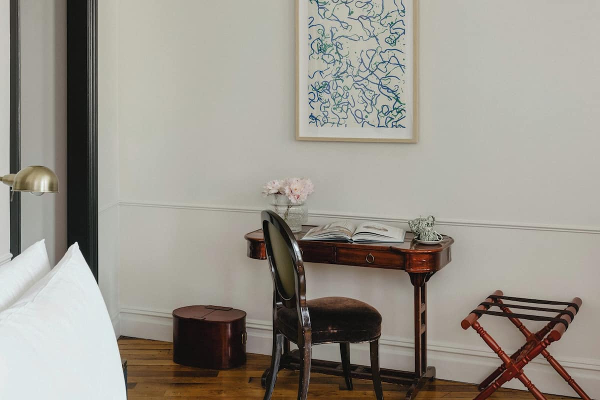 A hotel room with a bed, a desk, a chair, a small table with a vase, and a framed abstract artwork on the wall.
