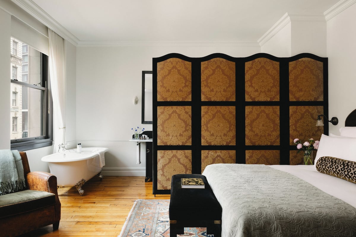 A bedroom with a large bed, a vanity with a mirror, a bathtub, and a view of the city through a window.