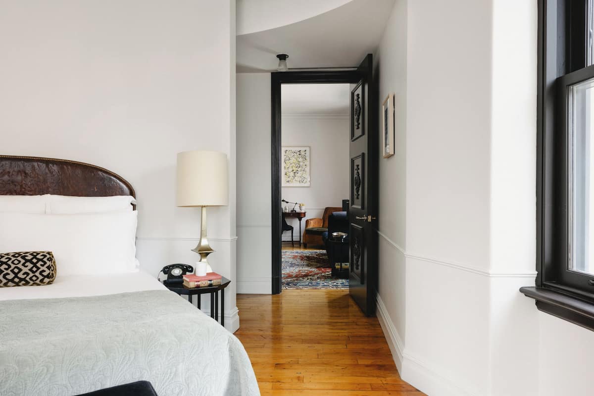 A hotel room with a bed, bedside table, lamp, and a view of the hallway.