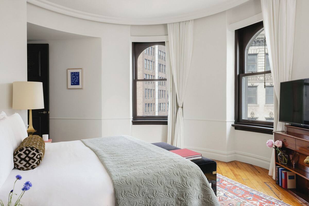 Una camera d'hotel con un letto grande, una televisione, una scrivania con lampada, una libreria e due finestre con tende.