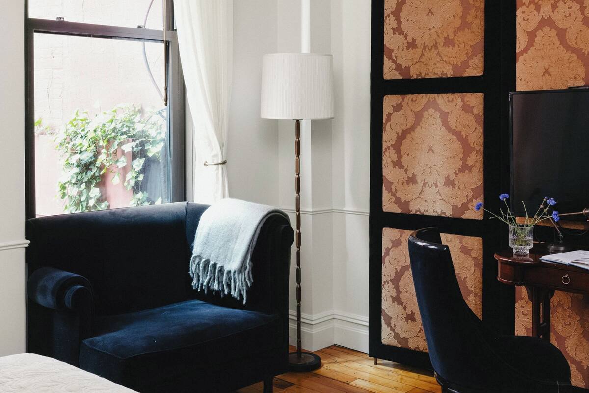 A room with a dark blue sofa, a white lamp, a desk with a chair, and a large window with curtains.