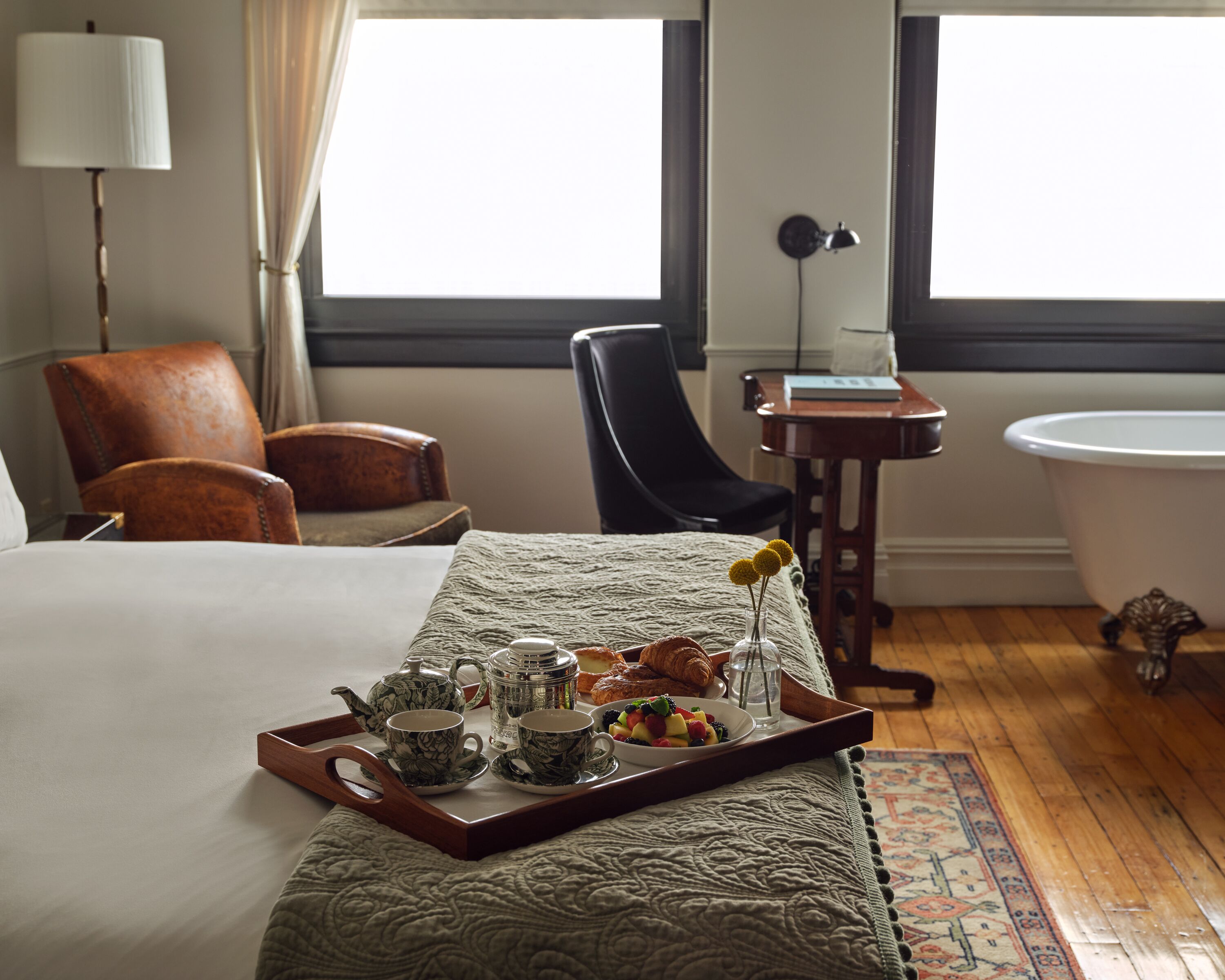 Une chambre d’hôtel avec un lit, une table de chevet en bois, une chaise, une lampe, une salle de bain avec une baignoire et un plateau avec des articles pour le petit-déjeuner.