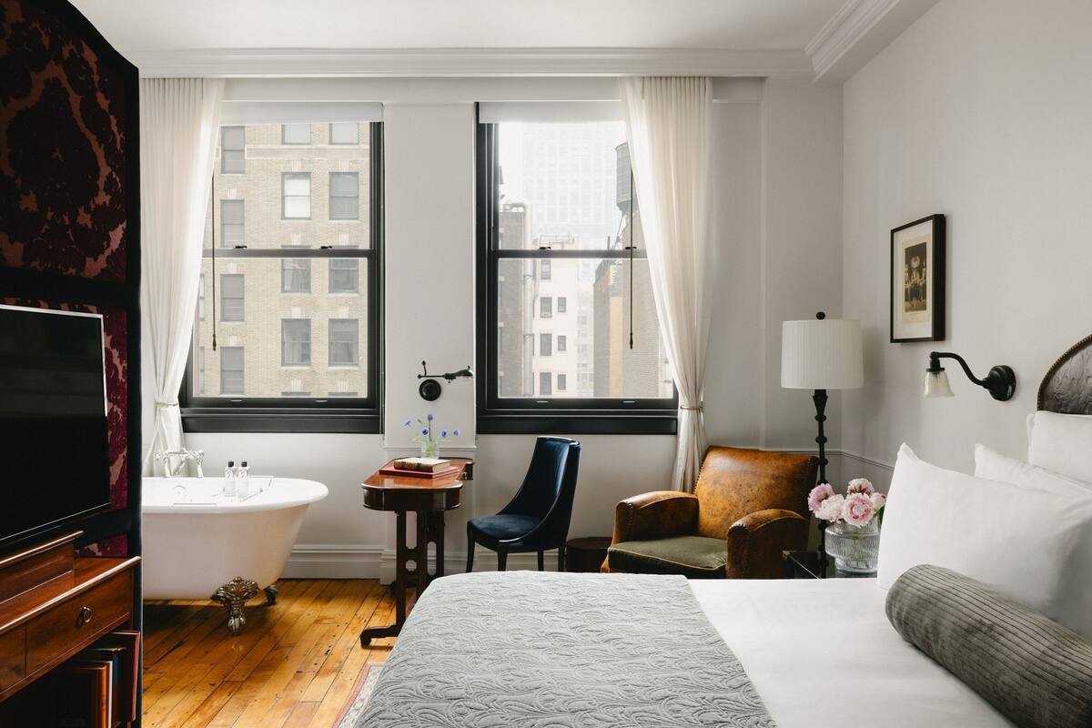 Una camera d'albergo con un ampio letto, un'area salotto con una poltrona e un tavolo, una vasca da bagno e vista sugli edifici attraverso ampie finestre.