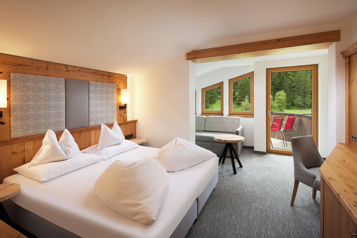 A hotel room with a large bed, a wooden headboard, a small table, a chair, and a balcony with a view of trees.