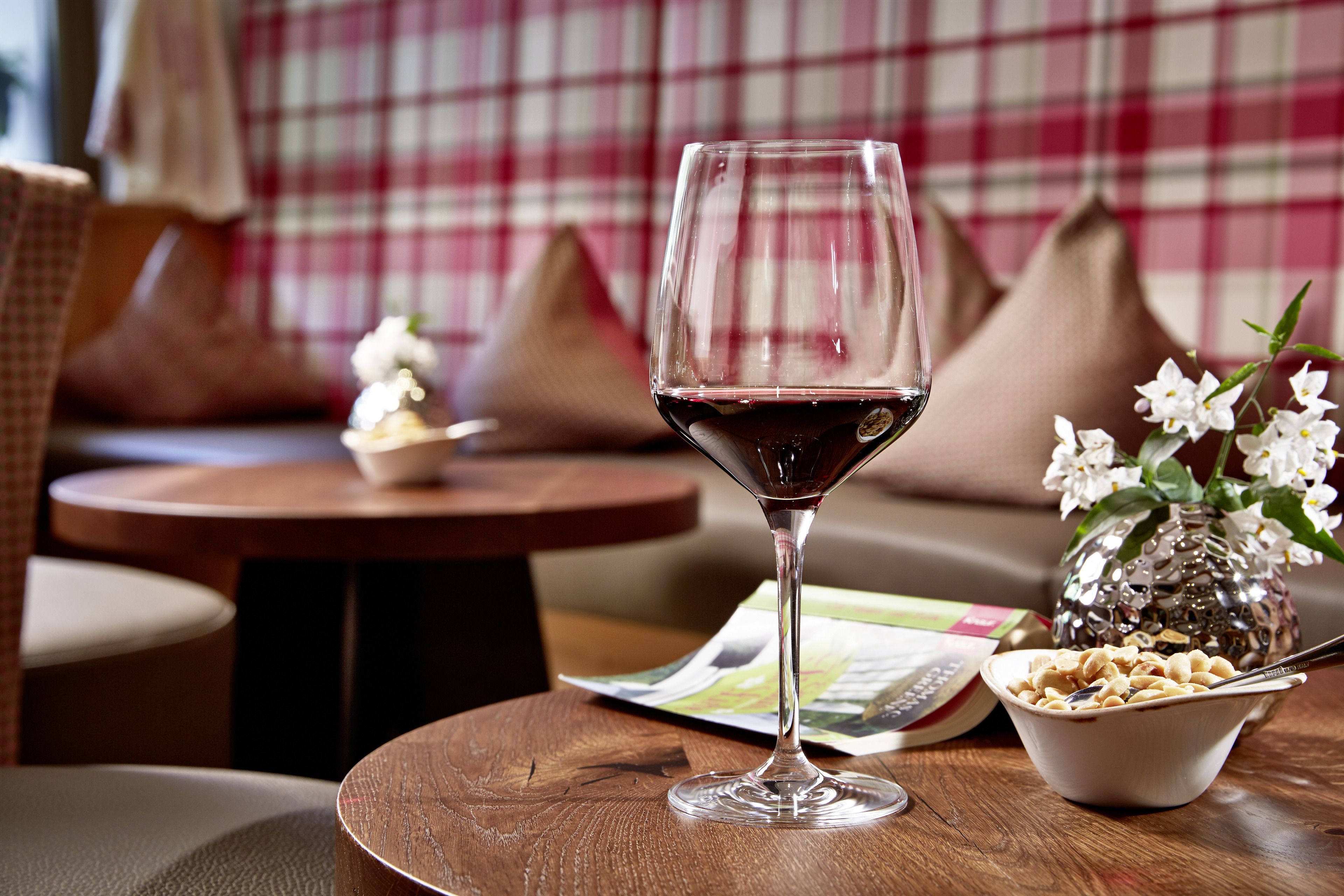 Ein Weinglas mit rotem Wein auf einem Holztisch, eine Schüssel mit Frühstückscerealien, eine Zeitschrift und eine Vase mit Blumen.