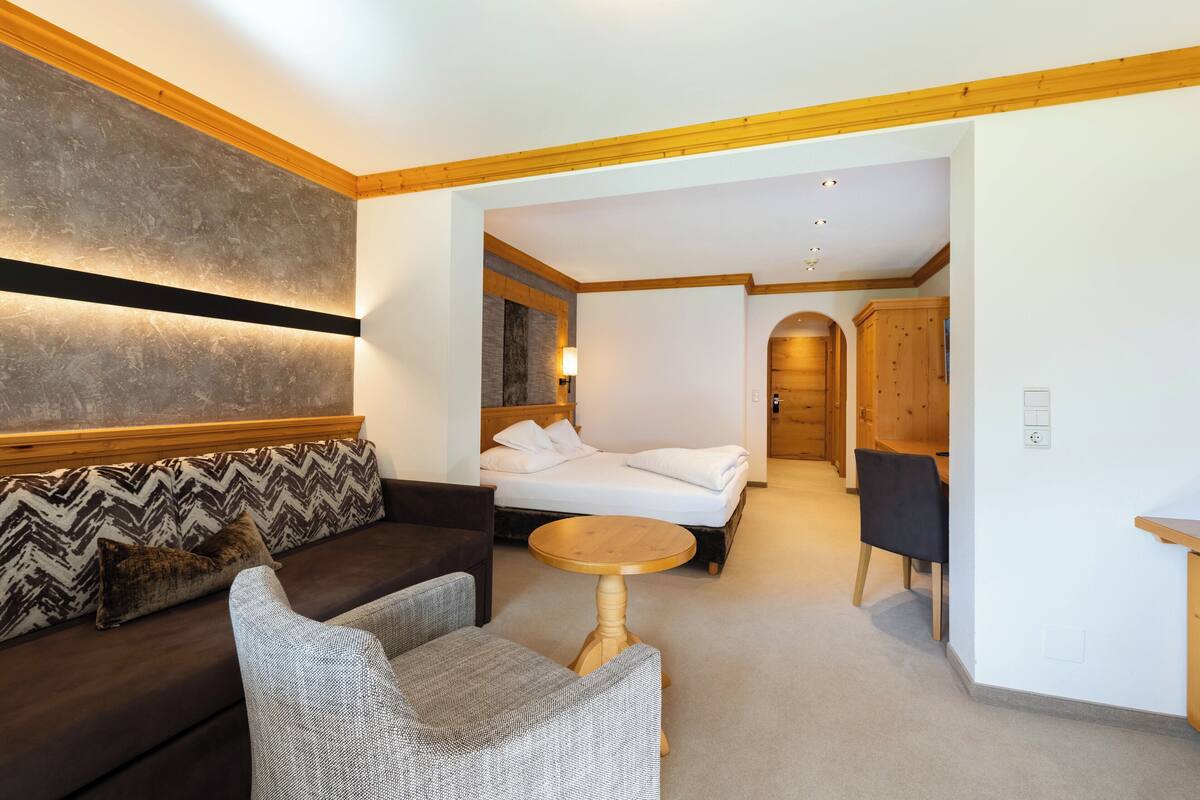 A modern hotel room with a sofa, a round wooden table, a bed, and a wooden cabinet.