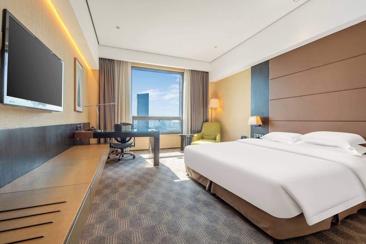 A hotel room with a large bed, a desk with a chair, a TV mounted on the wall, and a window with a city view.