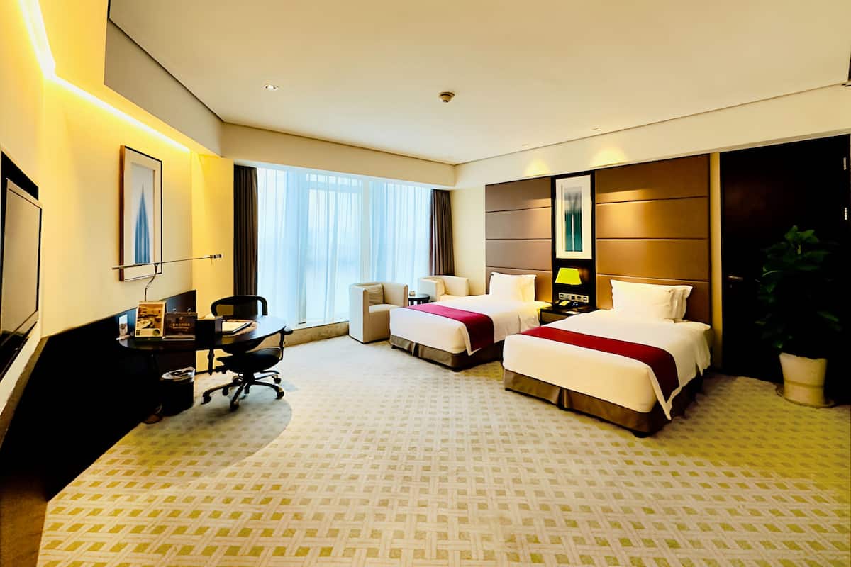 A hotel room with two beds, a desk, a chair, a lamp, and a large window with curtains.